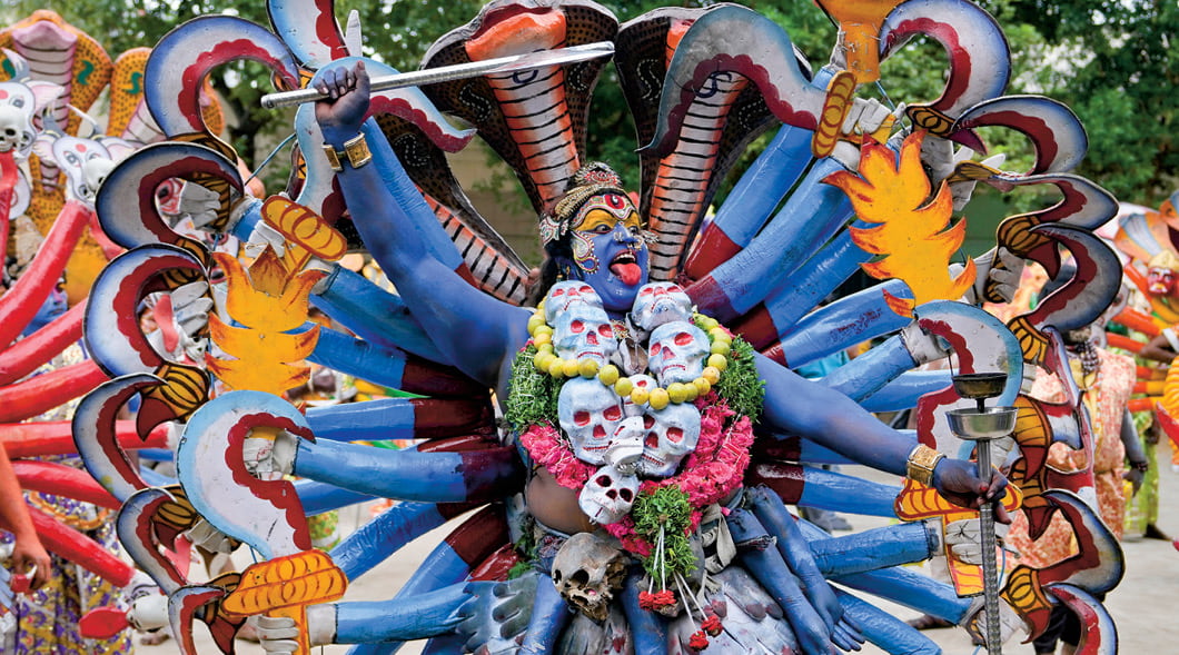 Image of a person painted blue and having many different arms while wearing skulls as a necklace