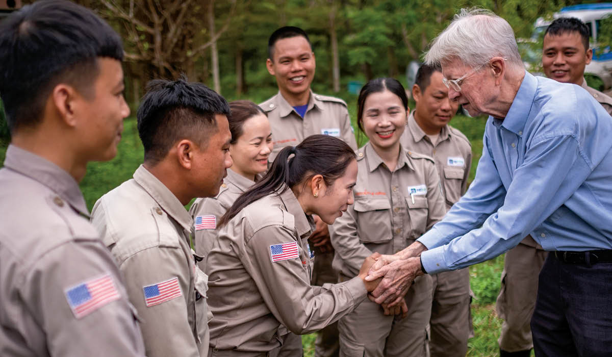 Photo of an older white man shaking the hands of Vietnamese people wearing American flag on uniforms