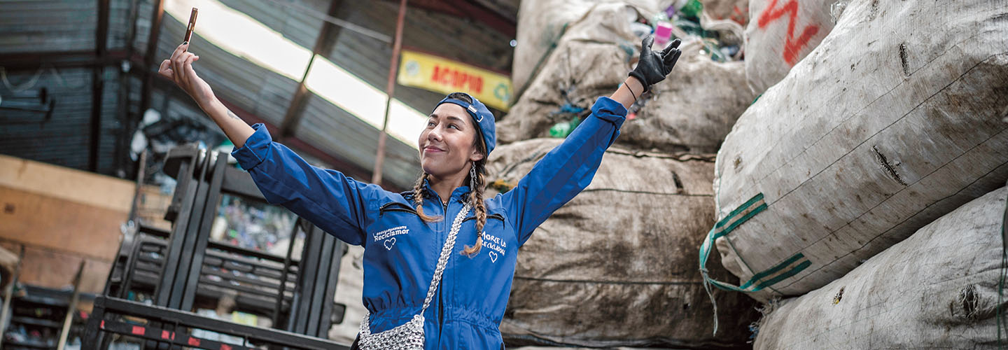 A woman in blue work uniform raises her arms in celebration beside large recycling bags