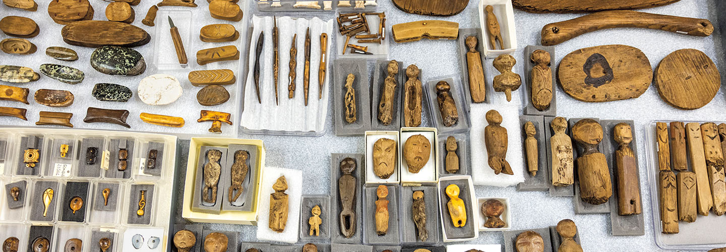 A collection of ancient carved wooden figures, tools, and stone artifacts arranged in trays
