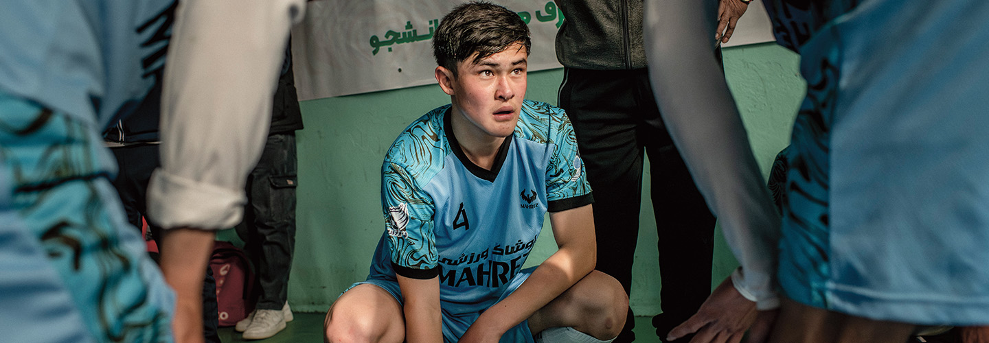 A young soccer player in a number 4 jersey crouches among teammates on a sideline bench