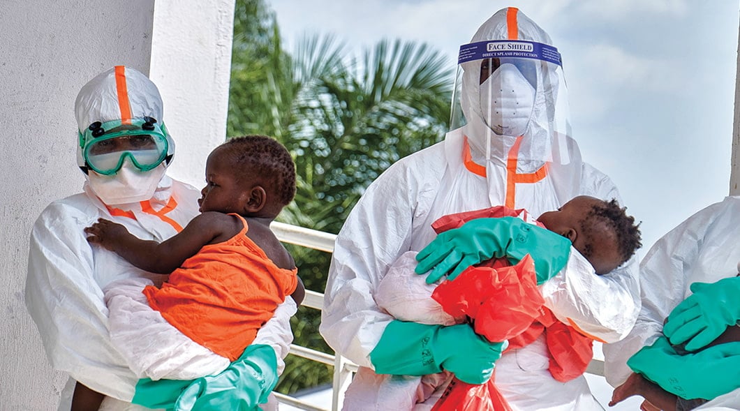 Two health workers in full protective gear and face shields each hold a young child