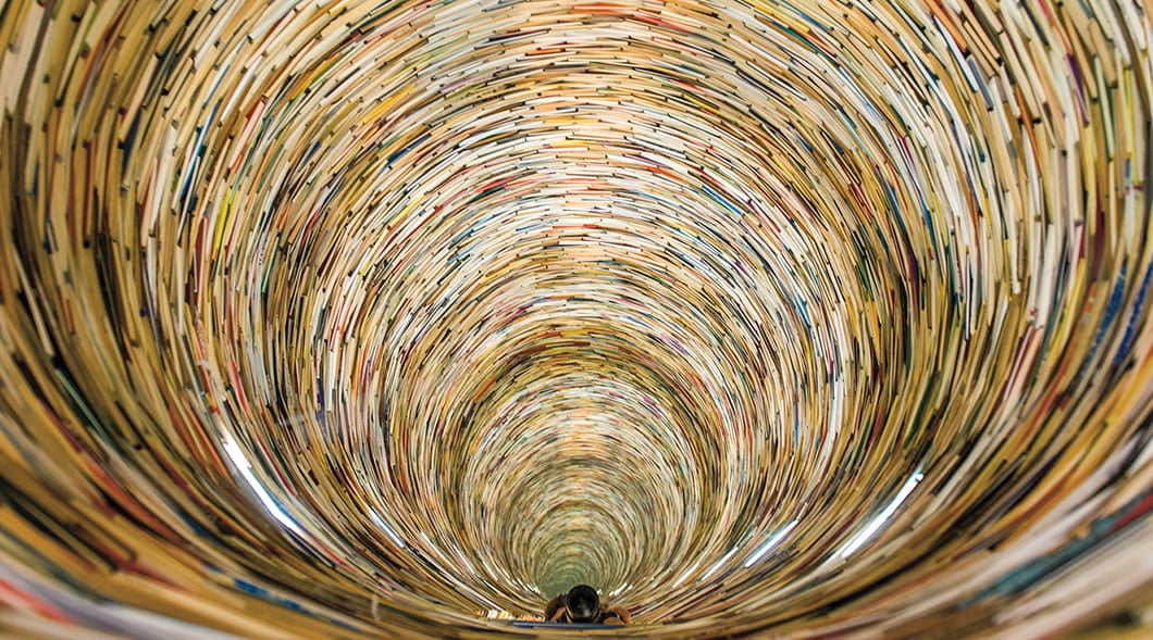 A person stands inside a towering cylindrical tower built from thousands of stacked books