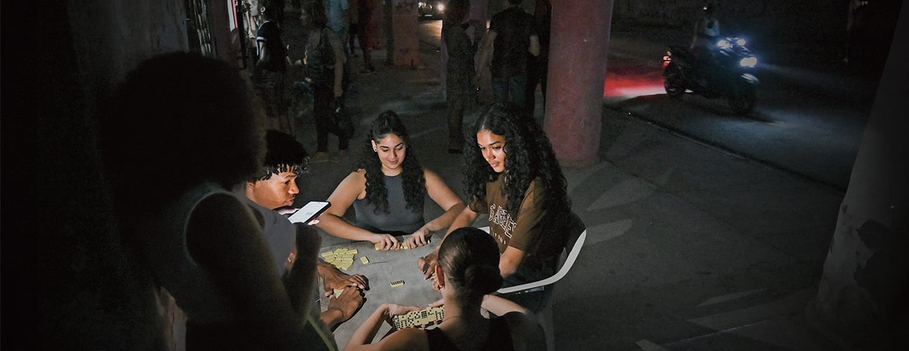 Four young people play dominoes around a table on a dark street at night while a motorcycle passes