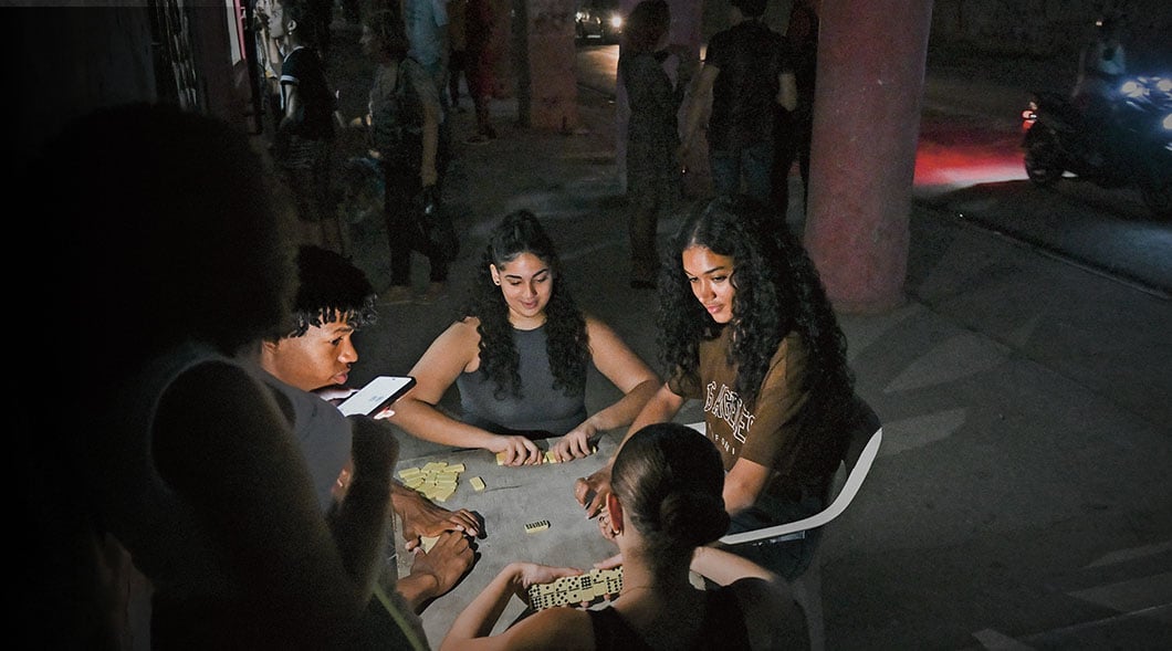 Young people play dominoes at a small table on a dark street while others stand nearby at night