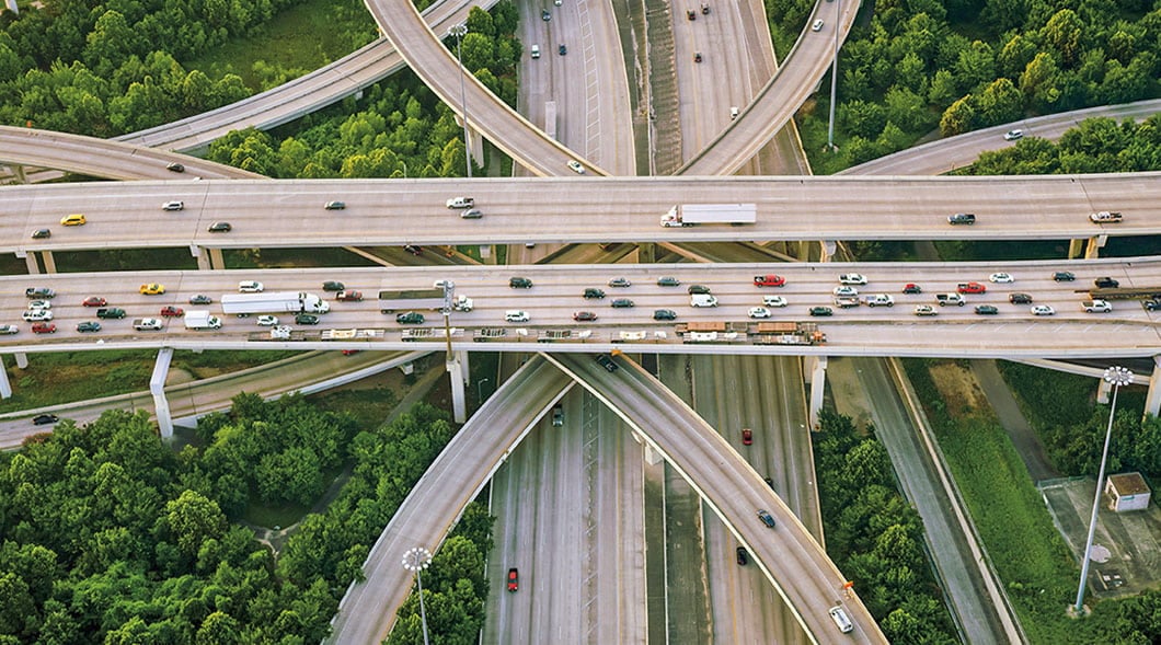 Cars & trucks cross stacked highway ramps at a wide interchange ringed by dense green trees