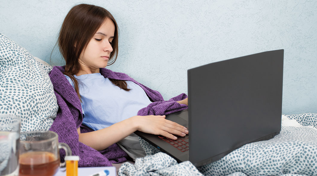A young woman in a purple blanket types on a laptop in bed beside a mug of tea & a pill bottle
